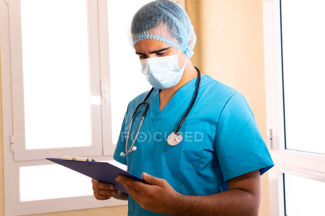 Médico masculino focado com estetoscópio e máscara lendo relatório médico na área de transferência enquanto estava na clínica — Fotografia de Stock