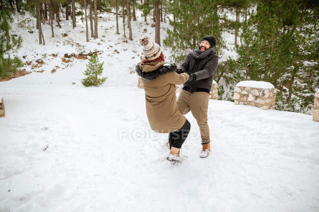 Entzücktes Paar in warmer Kleidung hält Händchen und dreht im verschneiten Winterwald herum, während es Spaß hat — Stockfoto