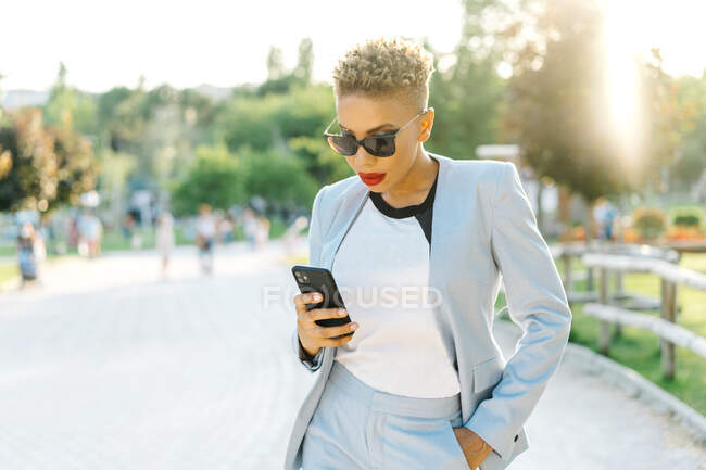 Elegante donna afroamericana in occhiali da sole messaggistica di testo sul cellulare mentre passeggia sulla passerella nel parco urbano — Foto stock