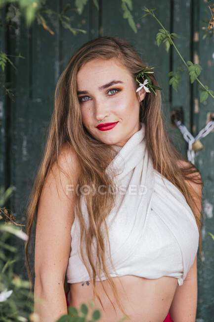 Delizioso femminile delicato con labbra rosse e fiori nei capelli lunghi guardando la fotocamera mentre in piedi in patio di casa in estate — Foto stock
