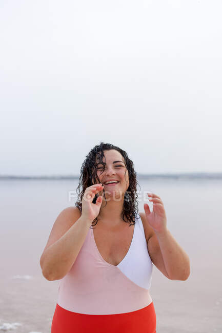 Mulher curvilínea positiva em maiô em pé na praia perto da lagoa rosa e olhando para a câmera enquanto desfruta de férias de verão — Fotografia de Stock