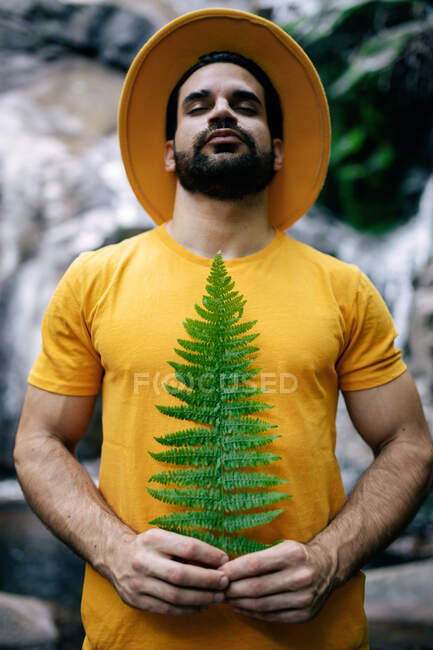 Спокійний чоловік мандрівник в жовтому одязі, що стоїть з зеленим листям папороті і насолоджується природою з закритими очима на тлі водоспаду в лісі — стокове фото