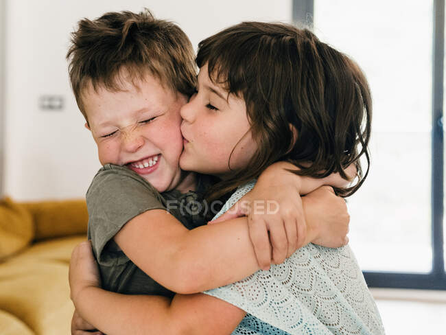 Lindos gemelos de contenido abrazando tiernamente y besándose juntos en la sala de estar en casa - foto de stock