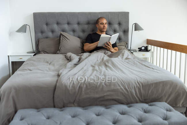 Проникливий самець сидить на м'якому ліжку вранці і читає цікаву історію в книзі після пробудження — стокове фото