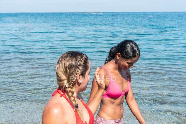 Content multiethnic female travelers in swimwear speaking on ocean beach against rocks during summer journey in sunlight — Stock Photo
