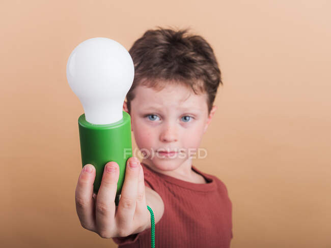 Pondering child in t shirt with plastic light bulb representing idea concept on beige background — Stock Photo