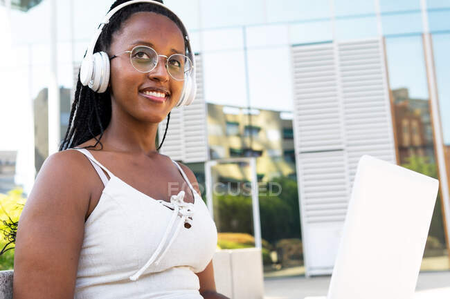 Mujer afroamericana positiva en auriculares sentada con portátil en la calle en Barcelona y disfrutando de canciones mientras mira hacia otro lado - foto de stock