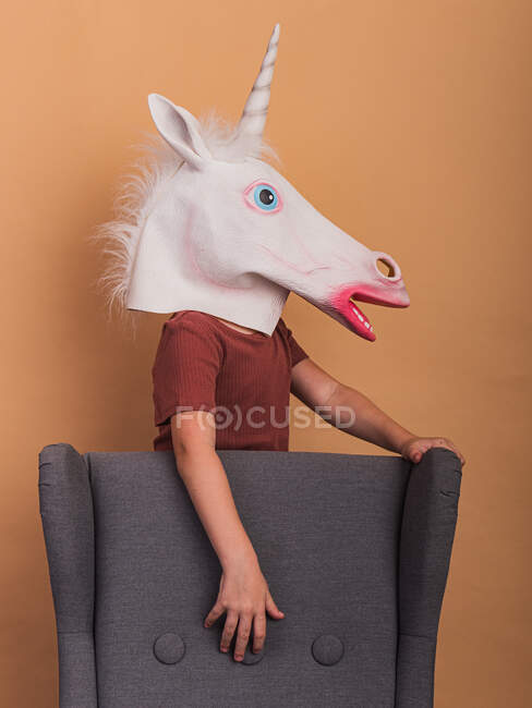 Вид збоку анонімної дитини в декоративній масці єдинорога з відкритим ротом, що торкається крісла на бежевому фоні — стокове фото