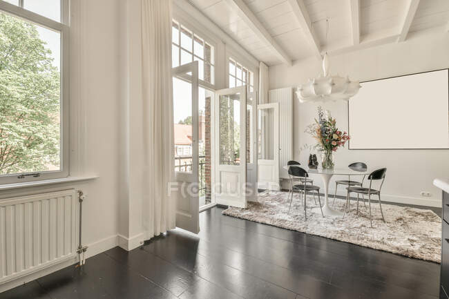 Moderno loft diseño interior de la casa con mesa de comedor y sillas colocadas cerca de la ventana en la esquina de la habitación espaciosa con imágenes simuladas colgando de la pared blanca en la casa - foto de stock