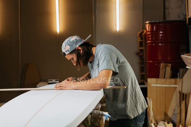 Vista lateral del modelador masculino utilizando la herramienta de medición mientras trabaja con la tabla de surf en el taller - foto de stock
