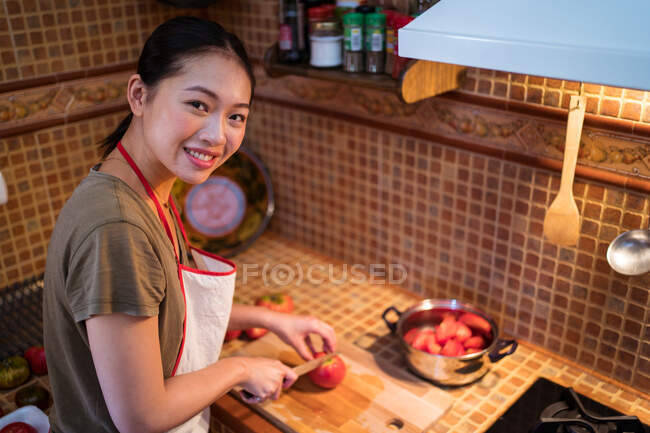 De cima étnica fêmea em avental de corte de tomates maduros em tábua de cortar enquanto cozinha almoço na cozinha em casa — Fotografia de Stock