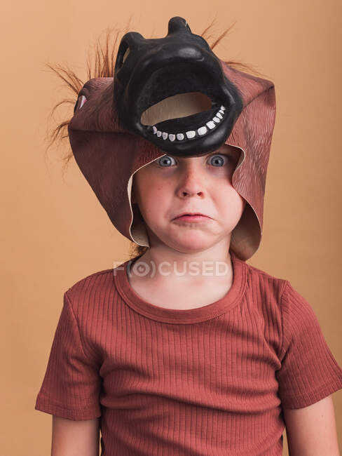Испуганный ребенок в футболке и маске лошади на голове смотрит в камеру на бежевом фоне — стоковое фото