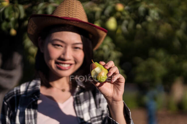 Delighted ethnic female farmer standing with ripe bitten pear in summer garden in countryside and looking at camera — Stock Photo