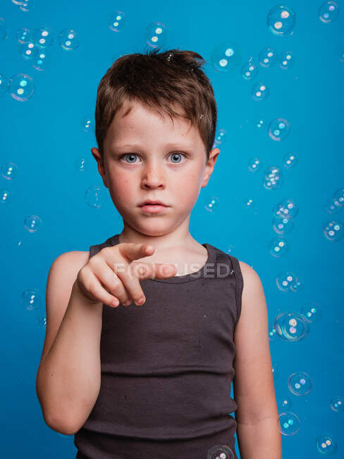 Étonné préadolescent garçon montrant geste touchant avec index dans l'air en studio avec bulles de savon volant sur fond bleu — Photo de stock