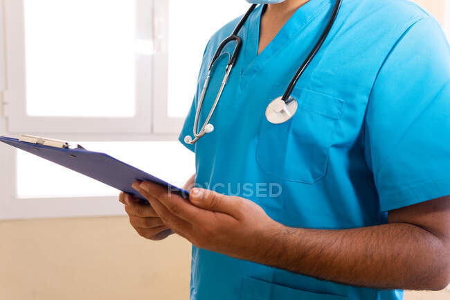 Medico maschio concentrato anonimo con stetoscopio e in maschera lettura rapporto medico sugli appunti mentre in piedi in clinica — Foto stock
