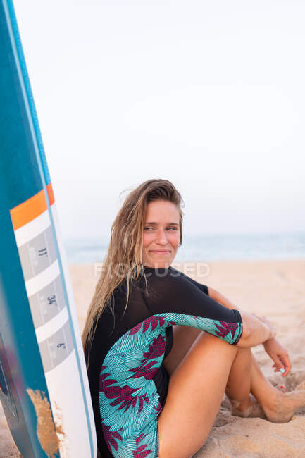 Alegre surfista femenina sentada con tabla SUP azul en la playa de arena en verano y mirando hacia otro lado - foto de stock