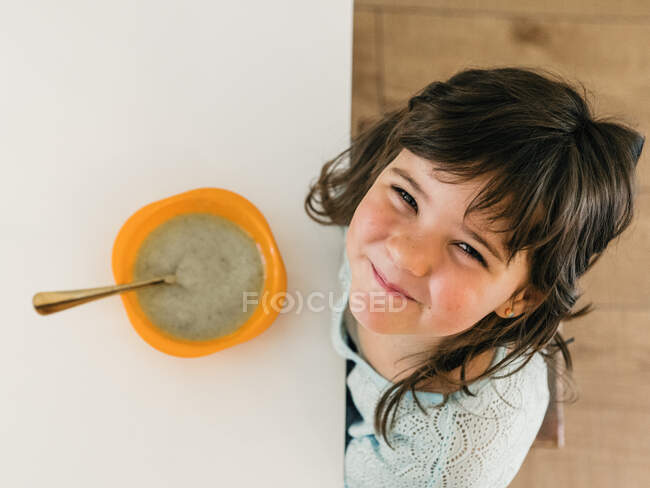 D'en haut de l'enfant joyeux assis à la table avec bol de soupe à la crème et regardant la caméra pendant le déjeuner dans la cuisine à la maison — Photo de stock