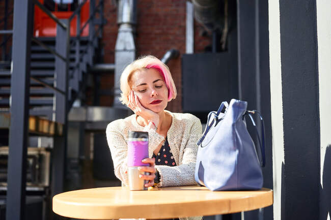 Спокойная неформальная женщина сидит за столом с экологически чистым кубком с горячим напитком в городе в солнечный день — стоковое фото