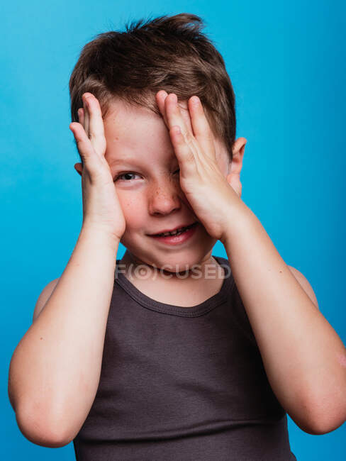 Schüchtern süß preteen boy cover eye mit hand und looking at camera on vid blue background im studio — Stockfoto