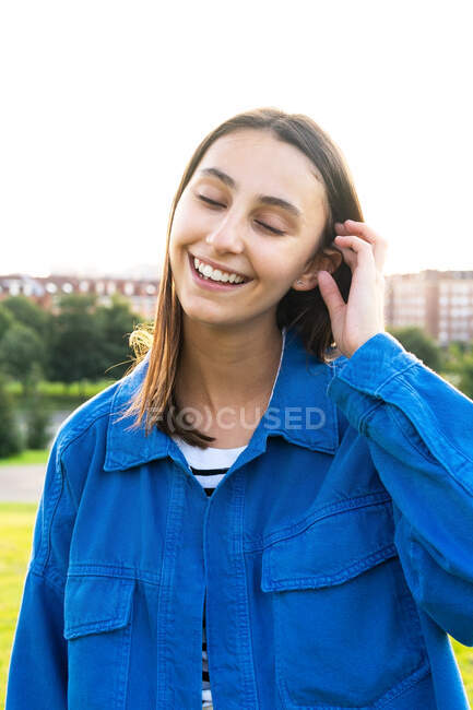Positive young female in trendy clothes standing with eyes closed smiling on green hill on background of city on sunny day — Stock Photo