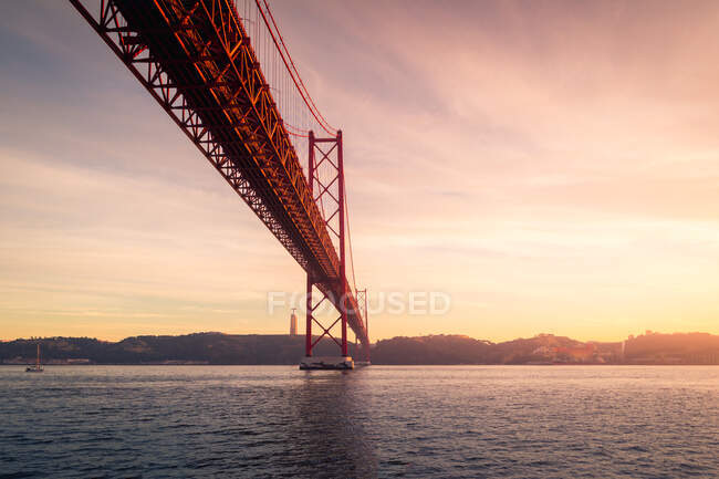 De baixo de postes de amarração de metal enferrujado localizados na margem do rio Tejo sob a Ponte 25 de Abril ao pôr-do-sol em Lisboa, Portugal — Fotografia de Stock