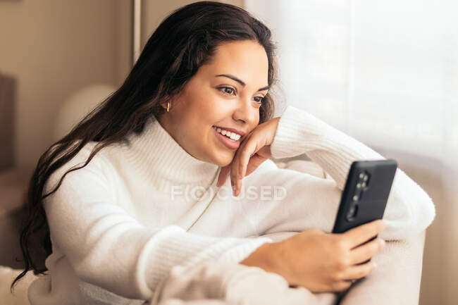 Весела жінка у светрі та джинсах сидить на дивані біля вікна і розмовляє на смартфоні. — стокове фото