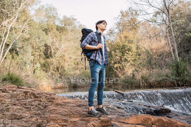 Escursionista maschio con zaino in piedi su un terreno roccioso vicino alla cascata nel bosco e guardando altrove — Foto stock