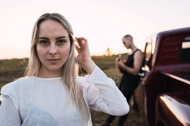 Ruhige Frau blickt in die Kamera vor dem Hintergrund eines verschwommenen Mannes, der in der Nähe eines Oldtimers in der Landschaft bei Sonnenuntergang steht — Stockfoto