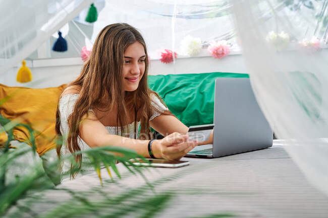 Mulher encantada sentada à mesa com laptop e fazendo compra com cartão de plástico durante compras on-line na tenda do quintal — Fotografia de Stock