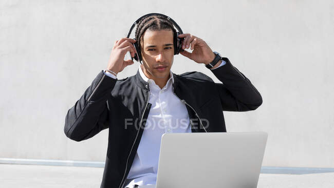 Jovem gerente masculino afro-americano com tranças em terno formal e fones de ouvido sem fio olhando para a câmera enquanto trabalhava em laptop na rua urbana — Fotografia de Stock