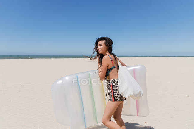 Vista lateral de la hembra positiva en traje de verano y con colchón inflable que camina en la costa arenosa en día soleado durante vacaciones - foto de stock
