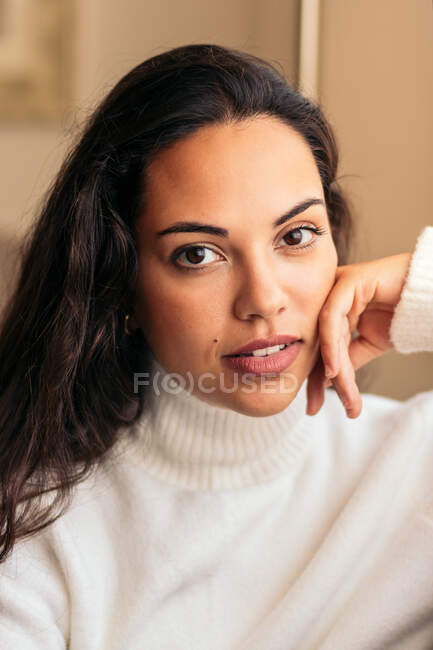 Женщина в белом свитере с рукой на лице смотрит в камеру рядом с окном — стоковое фото