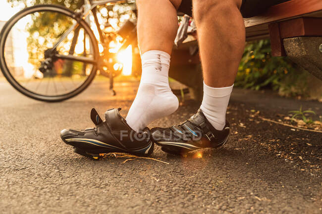 Crop unrecognizable male athlete in socks sitting on urban bench while taking off cycling shoes after training in sunlight — Stock Photo