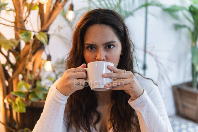 Jovem de cabelos longos latino-americano fêmea desfrutando delicioso café aromático de xícara de cerâmica, enquanto descansa em café acolhedor com plantas verdes — Fotografia de Stock