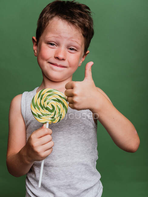 Delicioso niño preadolescente con caramelos remolinos mostrando como gesto mientras mira a la cámara en el fondo verde en el estudio - foto de stock