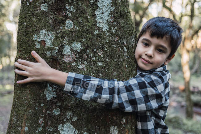 Amichevole bambino etnico in camicia a scacchi che abbraccia il tronco d'albero con muschio e licheni mentre guarda la fotocamera nella foresta — Foto stock