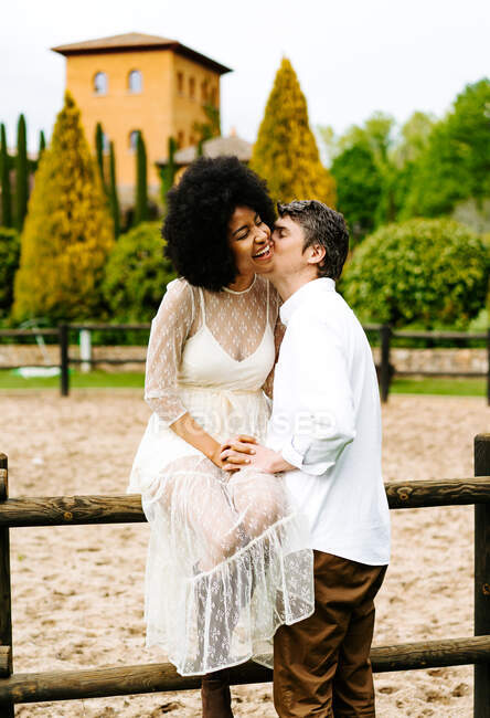 Tender man kissing content black woman sitting on wooden fence of paddock on ranch in summer — Stock Photo