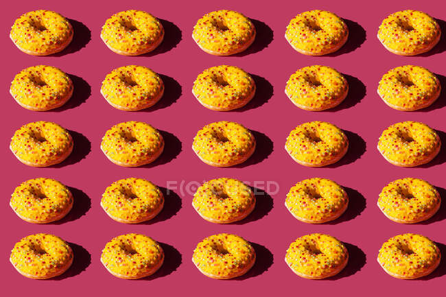 Вид зверху на багато пончиків, покритих жовтим покриттям і кольоровими кульками на рожевому фоні — стокове фото