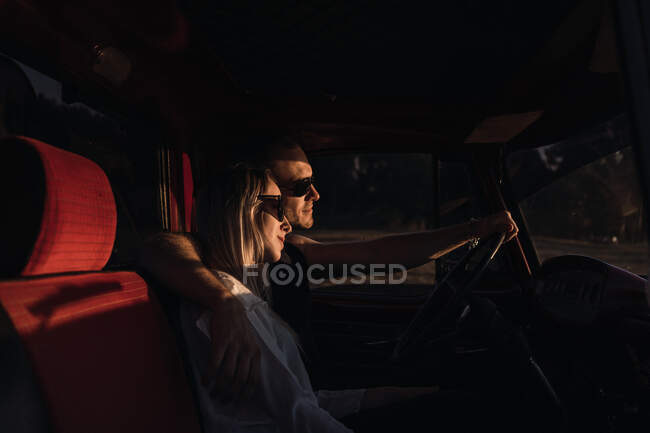 Side view of loving couple in sunglasses embracing in vintage car while riding in nature in evening — Stock Photo