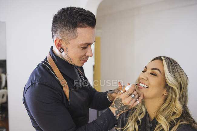 Cheerful blond woman smiling with closed eyes while tattooed makeup artist smearing liquid lipstick on lips of model — Stock Photo