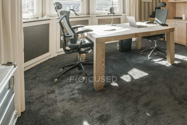 Table en bois avec ordinateur portable et fauteuils confortables placés près de la fenêtre panoramique dans une chambre spacieuse avec papier peint rayé dans un appartement contemporain — Photo de stock