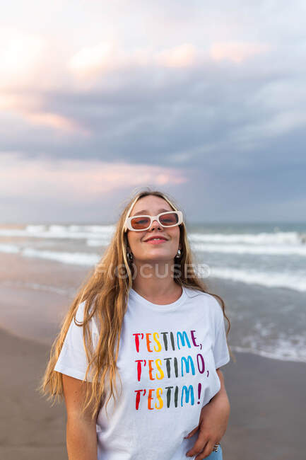 Mulher jovem positivo em óculos de sol na moda e roupa elegante em pé à beira-mar contra o mar na noite de verão — Fotografia de Stock
