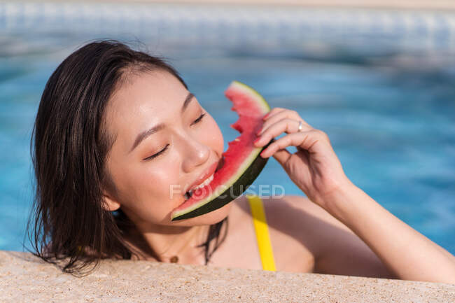 Vista lateral da alegre fêmea étnica em biquíni amarelo dentro da piscina e comer melancia fresca no dia ensolarado no verão — Fotografia de Stock
