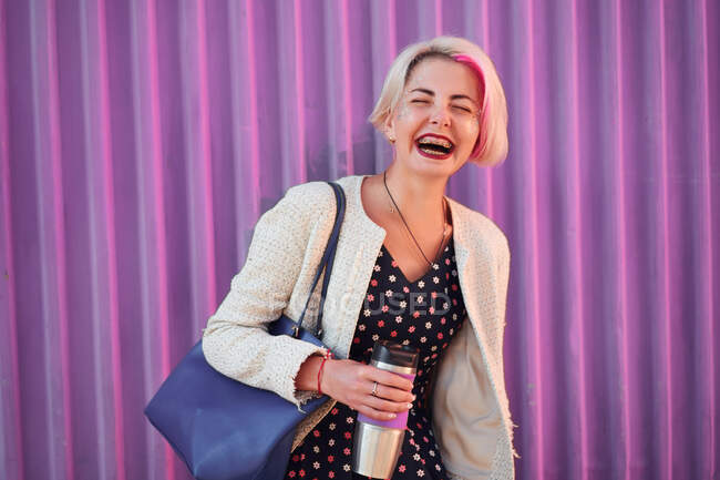 Femme informelle positive avec cheveux courts teints debout avec boisson dans une tasse écologique sur fond de mur violet en ville et les yeux fermés — Photo de stock