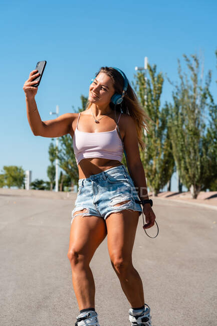 Patinadora femenina positiva en patines y auriculares que toma fotos en el teléfono móvil en un día soleado en verano en la ciudad - foto de stock