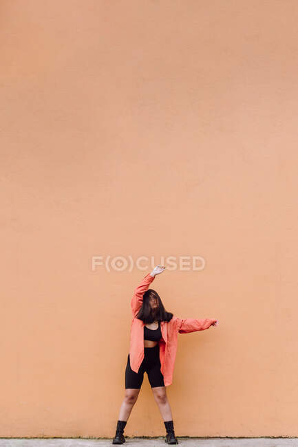 Cuerpo completo de joven mujer anónima cubriendo la cara con el pelo largo de color marrón levantando los brazos mientras está de pie contra la pared naranja - foto de stock