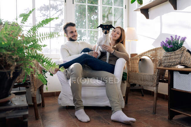 Uomo barbuto con fidanzata sorridente che abbraccia cane di razza mentre riposa in poltrona contro finestra nella stanza della casa — Foto stock
