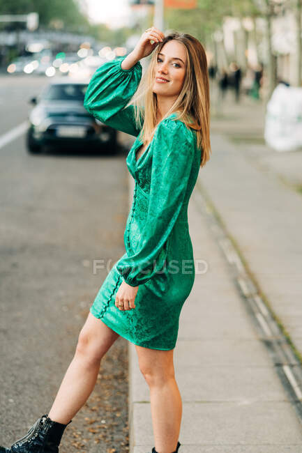 Mulher despreocupada em vestido verde na moda de pé com um braço levantado e tocando sua cabeça na rua e olhando para a câmera — Fotografia de Stock