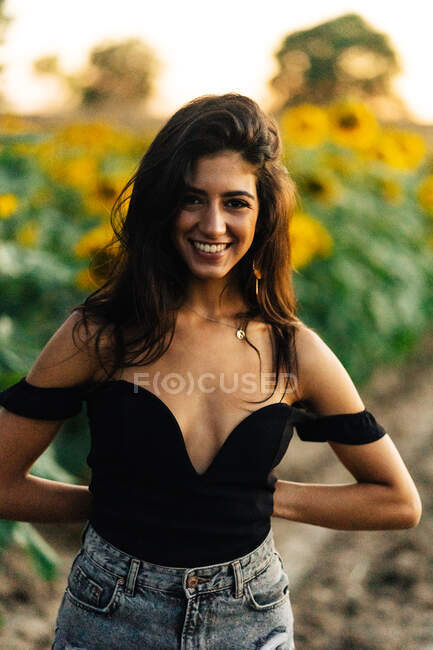 Charmante glückliche junge langhaarige hispanische Frau in schwarzem Top mit nackter Schulter, die neben blühenden gelben Sonnenblumen steht und an Sommertagen in der Natur in die Kamera schaut — Stockfoto