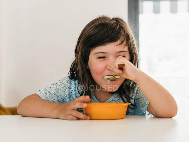 Criança adorável comendo deliciosa sopa de creme de tigela de plástico enquanto se senta à mesa durante o almoço em casa — Fotografia de Stock